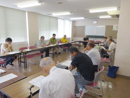 3階の研修室で行いました 2017年第2回定例会の様子。中央公民館3階の会議室。ブラインドが閉められていますが、外が明るいようで、隙間から光がたくさん見えます。斜め後ろからの写真で、写真の奥で事務局瀬戸が話しているのを13人が聞いています。一番手前には頭の薄くなった白いカッターシャツを着た人が座っています。